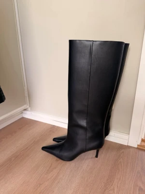 Svarta knähöga stövlar med klack från H&M - Säljer ett par svarta knähöga stövlar i slätt skinnimitation med spetsig tå och smal klack. Stövlarna har en stilren och elegant design som passar perfekt till höstens och vinterns outfits. Supersnygga till både jeans och kjol.