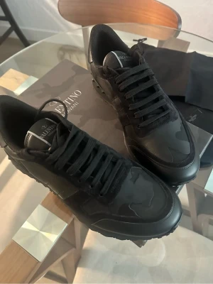 Svarta sneakers från Valentino - Snygga svarta sneakers från Valentino med diskret kamouflagemönster och svarta skosnören. Skorna har en klassisk låg siluett och är tillverkade i en mix av läder och textil. Perfekta för dig som gillar stilrena och exklusiva sneakers.