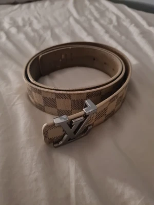 Beige rutigt bälte med silverdetalj - Snyggt bälte i beige och brunt med rutigt mönster och en stor silvrig spänne framtill. Perfekt accessoar för att lyfta din outfit med en exklusiv känsla. Bältet har klassisk design och passar till många olika stilar.