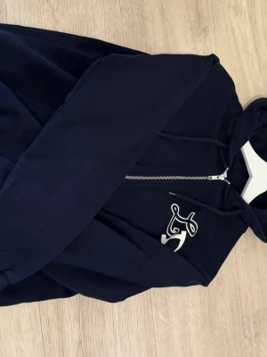 Mörkblå hoodie från Les Deux - Snygg mörkblå hoodie från Les Deux med dragkedja framtill och vit broderad logga på bröstet. Klassisk huva med dragsko och mjukt material som känns skönt mot huden. Perfekt för en avslappnad och stilren look.