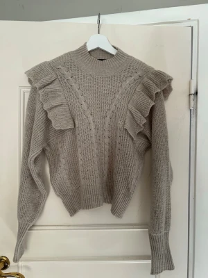 Beige stickad tröja med volanger - Supersnygg beige stickad tröja med volangdetaljer över axlarna och kabelstickat mönster framtill. Tröjan har hög hals, långa ärmar och ribbade muddar. Perfekt för dig som gillar trendiga detaljer och en mysig look.