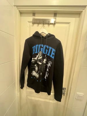 Svart hoodie med Biggie-tryck - Svart hoodie med stor blå text 'BIGGIE' och svartvitt tryck av Notorious B.I.G. på framsidan. Hoodien har dragsko i huvan och en klassisk känguruficka. Perfekt för dig som gillar streetwear och hiphopstil.