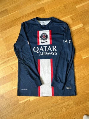PSG långärmad matchtröja blå - Snygg långärmad matchtröja från PSG i mörkblått med vita och röda detaljer. Tröjan har klubbmärke och Nike-logga på bröstet samt Qatar Airways tryckt framtill. Tillverkad i ett lätt och funktionellt material, perfekt för fotboll eller träning.