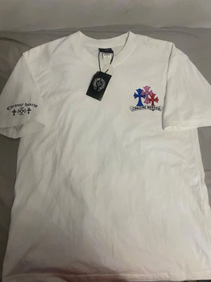 Vit Chrome Hearts t-shirt med tryck - Vit t-shirt från Chrome Hearts med färgglada kors i blått, rosa och rött på bröstet samt logotyp. Tryck på ena ärmen med text och små kors. Klassisk passform och rund halsringning. Perfekt för dig som gillar streetwear och detaljer.