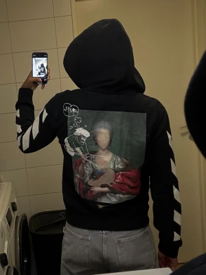 Svart Off-White hoodie med tryck - Svart hoodie från Off-White med dragkedja, vita pilar längs ärmarna och flera tryck framtill. Stor grafisk print på ryggen med konstmotiv och tecknad figur. Huva med snören och fickor fram. Perfekt för dig som gillar streetwear och unika detaljer.