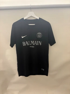 Svart PSG x Balmain t-shirt - Snygg svart t-shirt med PSG och Balmain Paris-logga framtill samt Nike-märke. Klassisk rund hals och korta ärmar. Perfekt för dig som gillar streetwear och sportig stil.