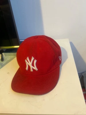Röd New Era Yankees keps - Snygg röd keps från New Era med klassisk New York Yankees-logga broderad i vitt framtill. Kepsen har böjd skärm och MLB-logga baktill. Tillverkad i ett tåligt tygmaterial och har en stilren, sportig look.
