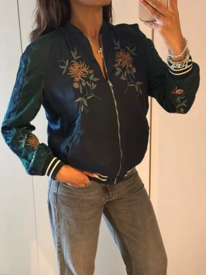 Mörkblå bomberjacka med blommor - Snygg mörkblå bomberjacka med broderade blommor i orange och grönt på bröstet och ärmarna. Jackan har dragkedja framtill, ribbade muddar med vita ränder och en avslappnad passform. Materialet ser ut att vara glansigt, troligen polyester eller satin.