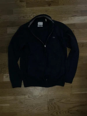 Mörkblå ziptröja från Gant - Mörkblå ziptröja från Gant med hög krage och diskret logga på bröstet. Tröjan har hel dragkedja framtill och ribbade muddar vid ärmslut och nederkant. Perfekt till lager på lager och enkel att matcha.
