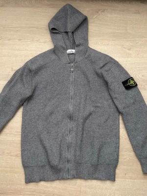 Grå stickad hoodie från Stone Island - Säljer en grå stickad hoodie från Stone Island med dragkedja framtill och ikonisk patch på vänster ärm. Hoodien har ribbade muddar och är tillverkad i ett mjukt, tjockt material som ger en schysst oversized look. Perfekt för dig som gillar streetwear och vill ha något bekvämt.
