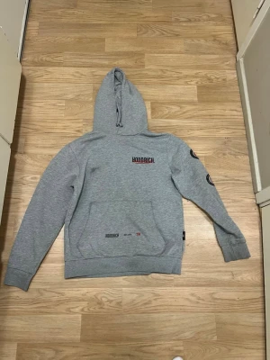 Grå hoodie från Hoodrich med tryck - Snygg grå hoodie från Hoodrich med svart och rött tryck på bröstet och ryggen. Hoodien har en stor huva, magficka och ribbade muddar. Det finns även tryck på ena ärmen som ger en streetwear vibe. Perfekt för dig som gillar avslappnad stil.