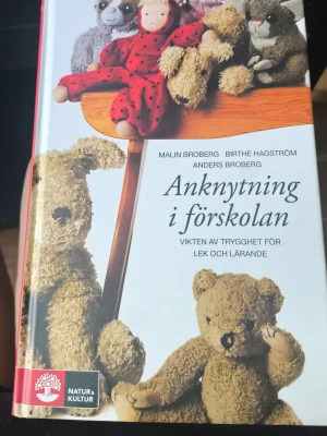 Anknytning i förskolan bok - Boken 'Anknytning i förskolan' av Malin Broberg, Birthe Hagström och Anders Broberg handlar om vikten av trygghet för lek och lärande i förskolan. Omslaget är vitt med bilder på gosedjur och en docka. Utgiven av Natur & Kultur.