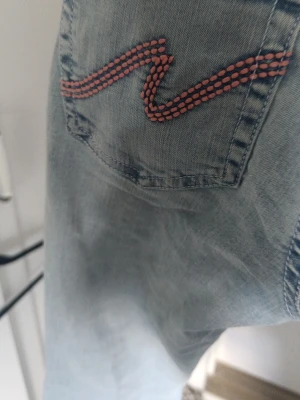 Ljusblå jeans med broderad ficka - Säljer ett par ljusblå jeans med en snygg broderad detalj i rosa på bakfickan och slitning vid knät. Jeansen har en avslappnad passform och är perfekta för en trendig och avslappnad stil. Materialet är klassisk denim i bomull.