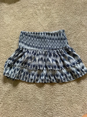 Blåvit volangkjol från Gina Tricot - Supersöt kort kjol från Gina Tricot i blått och vitt med smock i midjan och volangdetaljer. Kjolen har ett mönster som ger en fräsch och somrig känsla, perfekt till varmare dagar. Lätt och luftig modell som är enkel att matcha.