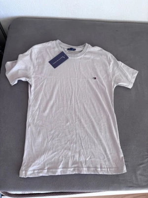 Vit t-shirt från Tommy Hilfiger - Klassisk vit t-shirt från Tommy Hilfiger med liten broderad logga på bröstet. T-shirten har rund halsringning och korta ärmar. Tillverkad i mjuk bomull för en skön känsla och enkel stil.