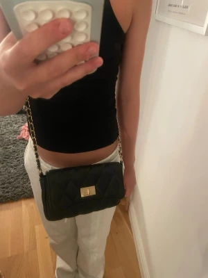 Svart quiltad handväska med kedja - Snygg svart handväska i quiltat mönster med guldfärgad kedjerem och en knapp för att stänga den . Väskan har en klassisk rektangulär form och är tillverkad i ett material som liknar konstläder. Perfekt accessoar för att lyfta din outfit.
