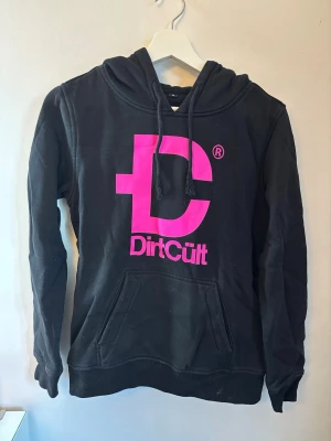 Svart hoodie med rosa DirtCult-tryck - Svart hoodie från DirtCult med stor rosa logga och text på bröstet. Klassisk känguruficka framtill, huva och ribbade muddar. Storleken är mer som en XS/S än en M. 