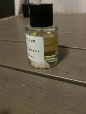 ESSNCE Drunk Apple Pie parfym - En stilren parfymflaska från ESSNCE med doften Drunk Apple Pie. Flaskan är cylindrisk i genomskinligt glas med svart lock och vit etikett med svart text. Innehållet är ljusgult och parfymen är en Eau de Parfum.