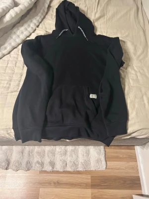 Svart hoodie med huva och magficka - En klassisk svart hoodie med stor huva och dragsnören. Hoodien har en rymlig magficka framtill och ribbade muddar vid ärmslut och nederkant. Perfekt för en avslappnad och stilren look. Materialet ser ut att vara mjukt och bekvämt, troligen bomullsblandning.