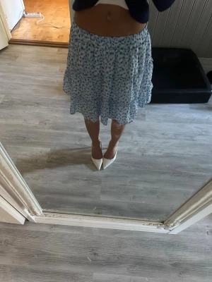 Blommig blåvit volangkjol - Söt kjol i vitt med blått blommönster och volangdetaljer. Kjolen har en luftig passform och resår i midjan, vilket gör den bekväm och enkel att bära. Perfekt för varma dagar och ger en fräsch sommarkänsla. Den är S men skulle säga att den både passar xs och nästan M med tanken på att den är stretchig