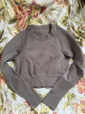 Långärmad och ribbad träningströja - Snygg grå croppad träningstopp med långa ärmar och ribbad struktur. Toppen har rund halsringning och är i polyamid och elastan. Sitter tajt och snyggt för en clean träningslook. Sparsamt använd.