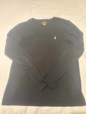 Svart långärmad topp från Polo Ralph Lauren - Klassisk svart långärmad topp från Polo Ralph Lauren med rund halsringning och den ikoniska broderade loggan på bröstet. Tillverkad i mjuk bomull som känns skön mot huden. Perfekt till jeans eller kjol för en clean och stilren look.