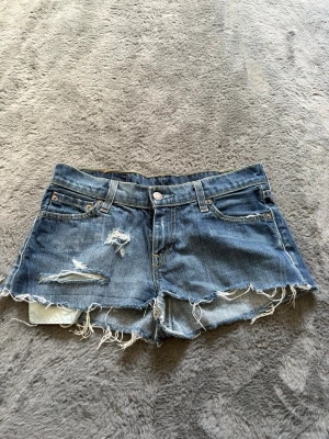 Blå slitna jeansshorts Levi's - Säljer ett par blå jeansshorts från Levi's med råa, fransiga kanter och slitna detaljer framtill. Klassisk femficksmodell med knapp och dragkedja. Perfekta för sommaren och ger en avslappnad vibe.