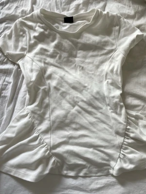 Vit peplum topp - Säljer en vit peplum topp med korta ärmar och rund hals. Toppen har en figurnära passform upptill och volang nedtill som ger en snygg siluett. Tillverkad i mjukt bomullsmaterial, perfekt för att styla med kjol eller jeans. Använd en gång innan