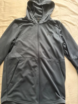 Mörkgrå hoodie från Under Armour - Snygg mörkgrå hoodie från Under Armour med dragkedja hela vägen och två sidofickor. Tillverkad i ett mjukt och stretchigt material som känns sportigt och bekvämt. Diskret logga på bröstet och klassisk huva för extra stilpoäng.