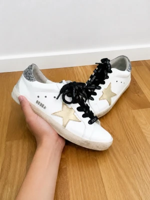 Golden Goose - Snygga vita sneakers med guldstjärna på sidan och silverglittrig häl. Skorna har svarta skosnören med text och en klassisk låg siluett. Perfekta för dig som vill sticka ut med detaljer och cool design.