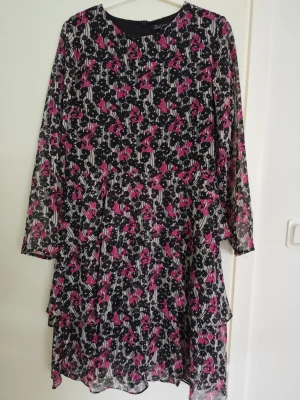Blommig svart och rosa kortklänning - Säljer en kort klänning med svart botten och blommigt mönster i rosa och vitt. Klänningen har långärmade, transparenta ärmar och volangdetaljer i kjolen. Tillverkad i ett lätt och luftigt material, perfekt för vår och sommar.