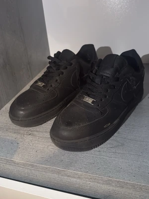 Nike air force 1 - Klassiska helsvarta Nike Air Force 1 sneakers i läder med perforerad tå, svarta skosnören och diskret Nike-logga på sidan och hälen. Snygg och tidlös modell som passar till det mesta. Metallbricka med 'AF1' på snörningen ger extra detalj.