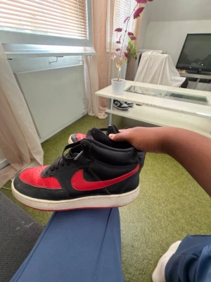 Svarta och röda high nike sneakers - Snygga high top sneakers i svart och rött med klassisk design. Skorna har svart ovandel med röda detaljer på tå, sidor och häl samt svart snörning. Yttersulan är vit med röd undersida. Perfekta för dig som vill sticka ut med en sportig look.