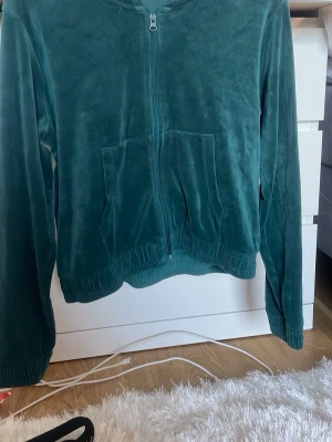 Grön velourhoodie med dragkedja - Mysig grön hoodie i mjuk velour med dragkedja framtill och två stora fickor. Tröjan har huva och ribbade muddar vid ärmslut och nederkant. Perfekt för en chill och avslappnad stil.