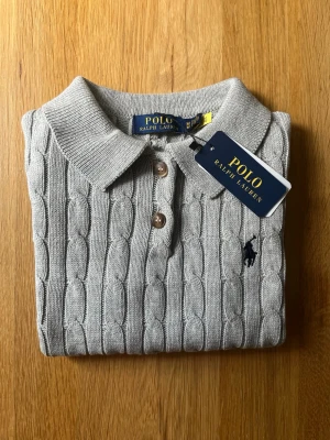 Grå kabelstickad skjorta Polo Ralph Lauren - Snygg grå kabelstickad skjorta från Polo Ralph Lauren med klassisk krage och knappar framtill. Skjortan har ett broderat mörkblått logotyp på bröstet och är tillverkad i mjukt material som ger en exklusiv känsla.