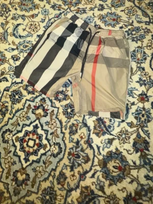 Randiga shorts med snörning - Säljer ett par snygga shorts med breda ränder i svart, beige, vitt och rött. Shortsen har elastisk midja med snörning och en avslappnad passform. Perfekta för sommaren och riktigt trendiga.