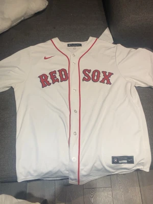 Vit Red Sox baseballtröja Nike - Säljer en vit baseballtröja från Nike med Red Sox-tryck i rött och svart på bröstet. Tröjan har knappar framtill, röd piping längs knappraden och korta ärmar. Perfekt för sport eller streetwear.