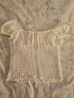 Vit topp från H&M - Söt vit topp från H&M med smockad midja och korta puffärmar. Toppen har en lätt rynkad överdel och är tillverkad i ett mjukt, tunt tyg som känns luftigt och skönt. Perfekt för en trendig och avslappnad look.