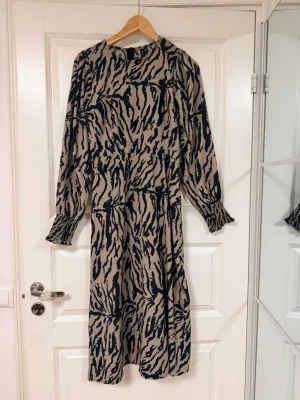 Beige och svart zebramönstrad klänning - Säljer en beige och svart långklänning med zebramönster. Klänningen har långa ärmar med smock vid ärmsluten och en avslappnad passform. Perfekt för dig som gillar djurmönster och vill ha något unikt i garderoben. Klänningen har väldigt skön material som passar perfekt för sommaren!
