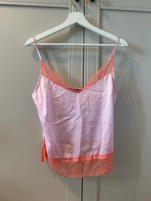 Ljusrosa satintopp med spetsdetaljer - Snyggt rosa linne i satin med tunna axelband och dekorativ rosa spets längs v-ringningen och nederkanten.