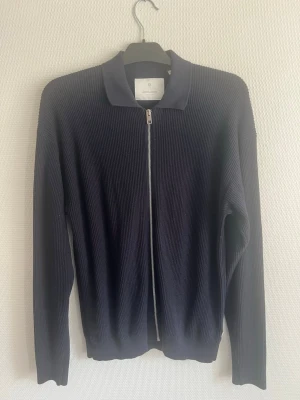 Marinblå ziptröja från Jack & Jones - Snygg marinblå ziptröja från Jack & Jones med ribbad struktur och klassisk krage. Tröjan har hel dragkedja framtill och långa ärmar. Perfekt för lager på lager och enkel att matcha med olika outfits.