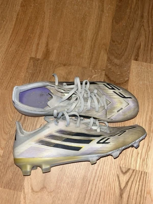 Adidas PRO F50 Road To Glory  - Säljer ett par vita fotbollsskor med svarta och gula detaljer. Skorna har snörning, låg profil och dobbar. Ovandelen är i syntetmaterial och sulan har text och mönster för extra grepp. Perfekta för dig som vill ha lätta och snabba skor på planen. Öppen för prisdiskutering!