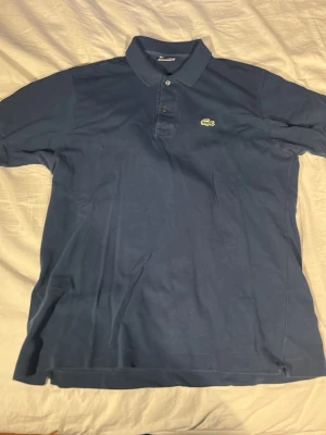 Blå piké från Lacoste - Säljer en klassisk blå pikétröja från Lacoste med korta ärmar och den ikoniska krokodilloggan broderad på bröstet. Tröjan har krage, knappar framtill och är gjord i bomull för en skön känsla. Perfekt för dig som gillar stilrena och tidlösa plagg.