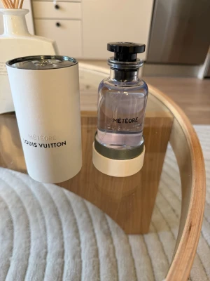 Louis Vuitton Météore parfym - Exklusiv parfym från Louis Vuitton, Météore, i en elegant genomskinlig glasflaska med svart lock. Flaskan har en stilren design och kommer med en vit cylinderformad förpackning. Doften är lyxig och modern, perfekt för dig som vill sticka ut.