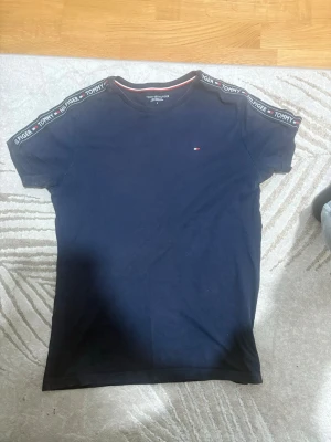 Mörkblå Tommy Hilfiger t-shirt - Snygg mörkblå t-shirt från Tommy Hilfiger med korta ärmar och diskret logga på bröstet. Axelbanden har ett band med Tommy Hilfiger-tryck för en sportig touch. Perfekt till jeans eller shorts.