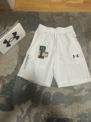 Vita träningsshorts från Under Armour - Snygga vita träningsshorts från Under Armour med elastisk midja och snörning. Loggan syns tydligt på ena benet och texten 'ARMOUR' i färgglada bokstäver på sidan. Perfekta för gymmet eller sport. Lätta och bekväma i syntetmaterial.