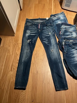 Mörkblå Dsquared2 slim jeans - Säljer ett par mörkblå slim fit jeans från Dsquared2 med slitningar, färgstänk och snygga detaljer. Jeansen har klassisk femficksmodell, guldfärgade sömmar och en röd Dsquared2-logga vid fickan. Perfekta för dig som gillar en trendig och avslappnad stil.