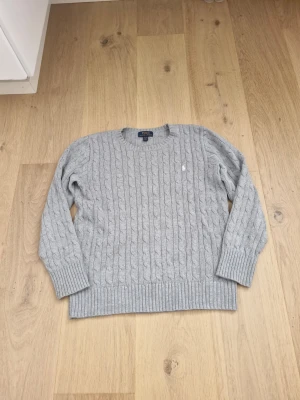 Ljusgrå kabelstickad tröja Polo Ralph Lauren - Kabelstickad tröja i ljusgrått från Polo Ralph Lauren. Klassisk rund halsringning, ribbade muddar och nederkant samt diskret broderad logga i vitt på bröstet. Perfekt för lager på lager och en stilren look. Hör av er vid frågor eller funderingar.