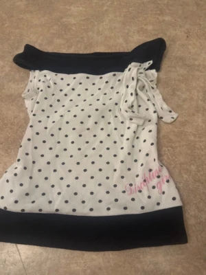 Vit och svart prickig offshoulder topp - Snygg offshoulder topp i vitt med svarta prickar och svarta kanter. Toppen har en knytning på ena axeln och rosa text. Perfekt för dig som gillar en lekfull och trendig stil.