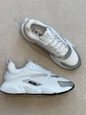 Vita och grå Dior sneakers - Chunky sneakers från Dior i vitt och ljusgrått med mesh och läderdetaljer. Skorna har en modern siluett, tjock sula och snygga detaljer i silver. Perfekta för dig som gillar streetwear och exklusiv design.